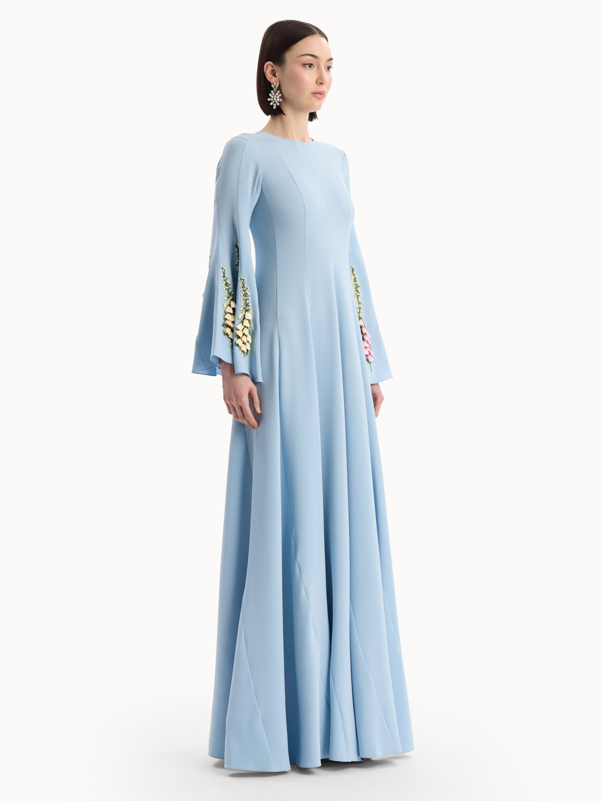 The model is wearing the Foxglove Embroidered Bell Sleeve Caftan in light blue featuring a floor-sweeping hemline and wide, bell sleeves embroidered with floral motifs. Side View.