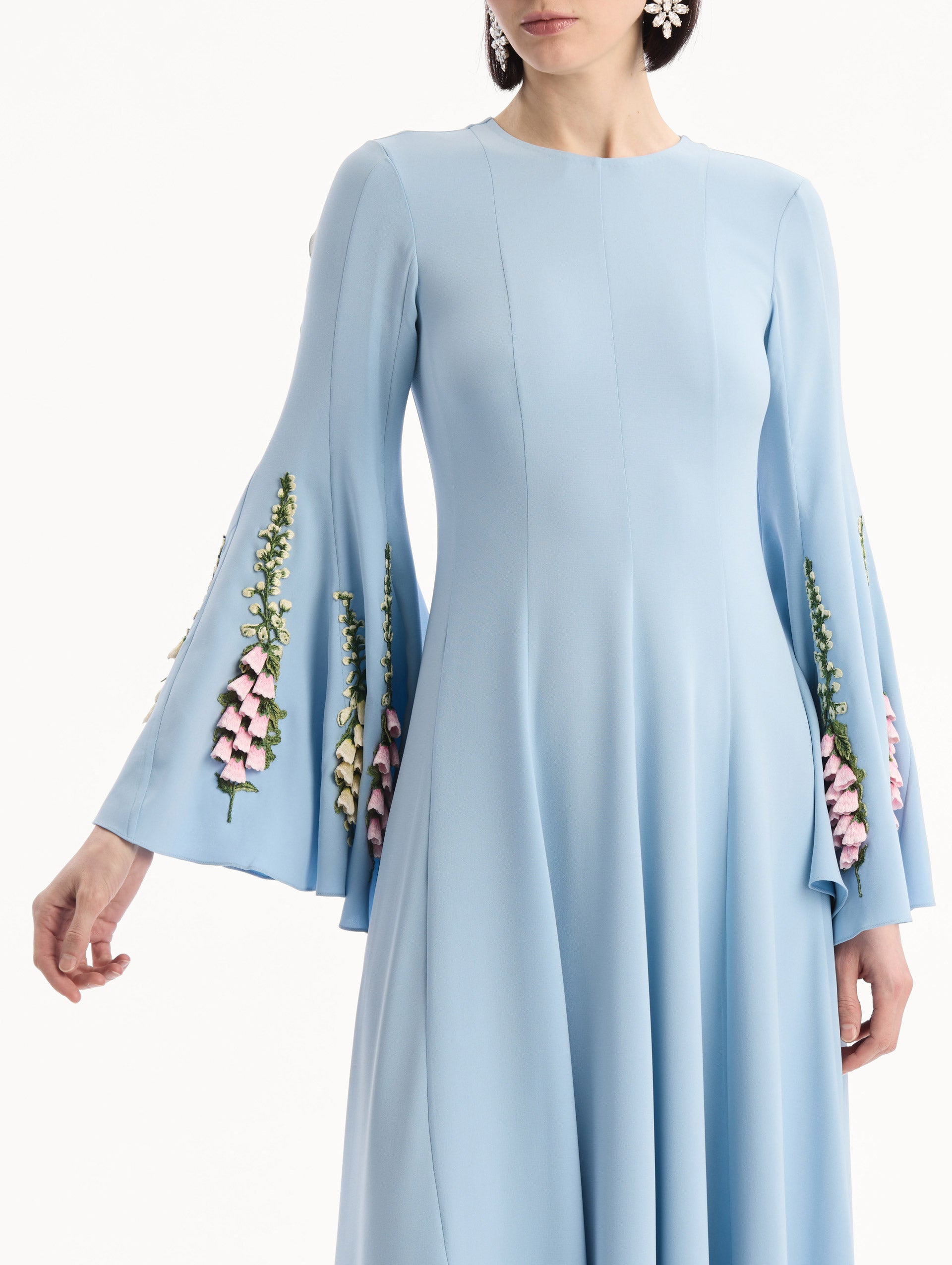 The model is wearing the Foxglove Embroidered Bell Sleeve Caftan in light blue featuring a floor-sweeping hemline and wide, bell sleeves embroidered with floral motifs. Front View.