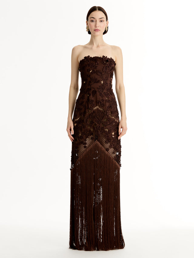 Mahogany strapless floor length gown with Porcelain flower threadwork in sculptural cutouts with glimpses of skin. Intricate lattice motifs. The silhouette elongates before cascading into a curtain of fringe. Front View.