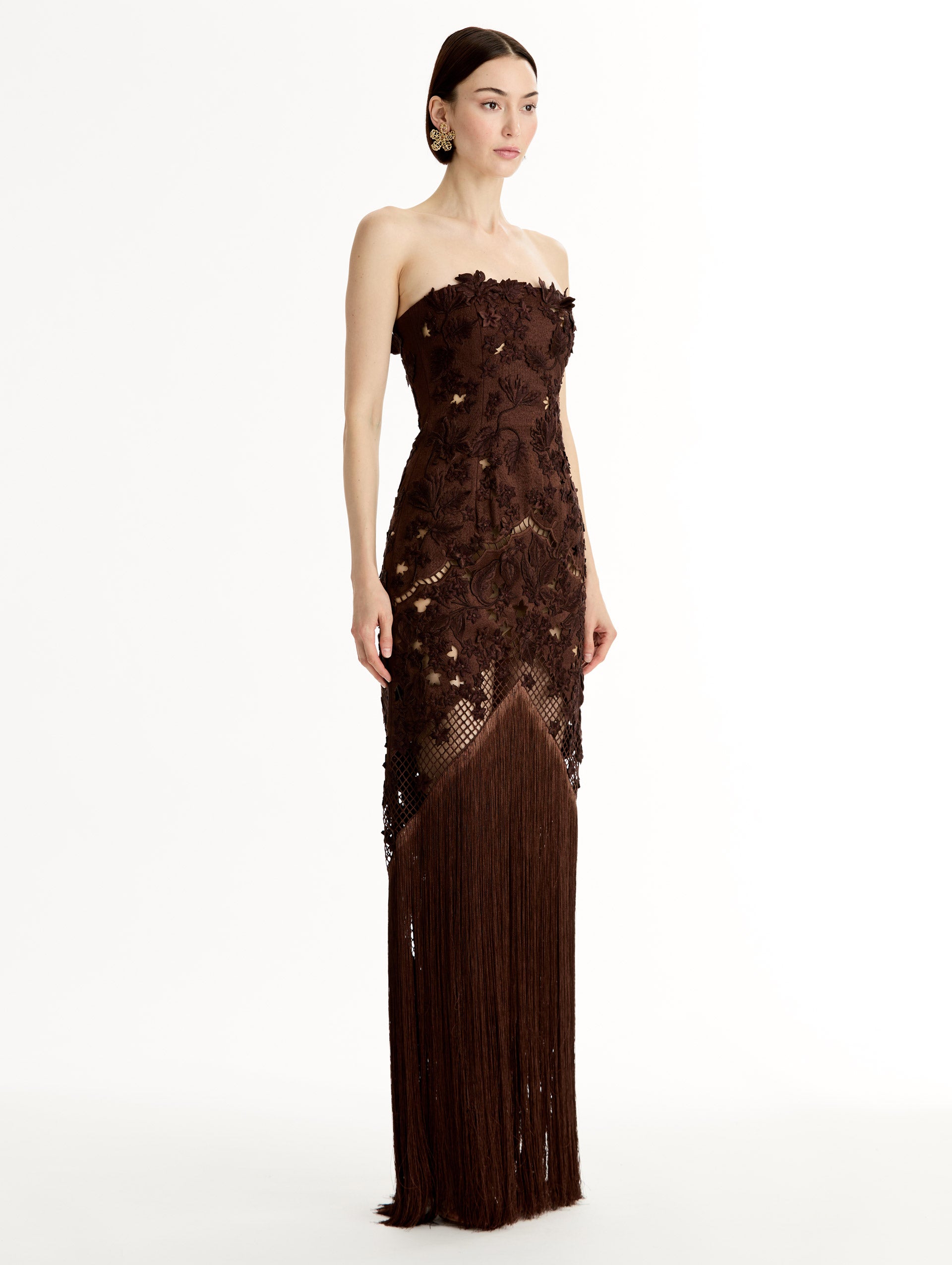 Mahogany strapless floor length gown with Porcelain flower threadwork in sculptural cutouts with glimpses of skin. Intricate lattice motifs. The silhouette elongates before cascading into a curtain of fringe. Side View.