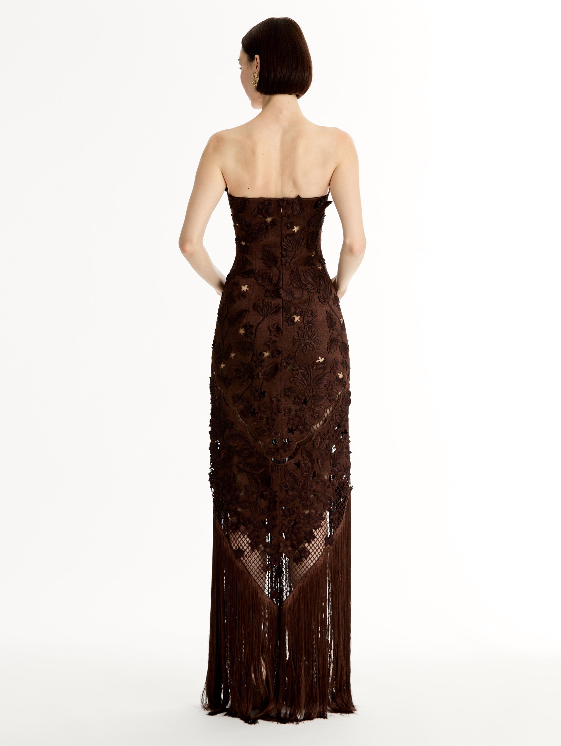 Mahogany strapless floor length gown with Porcelain flower threadwork in sculptural cutouts with glimpses of skin. Intricate lattice motifs. The silhouette elongates before cascading into a curtain of fringe. Back View.
