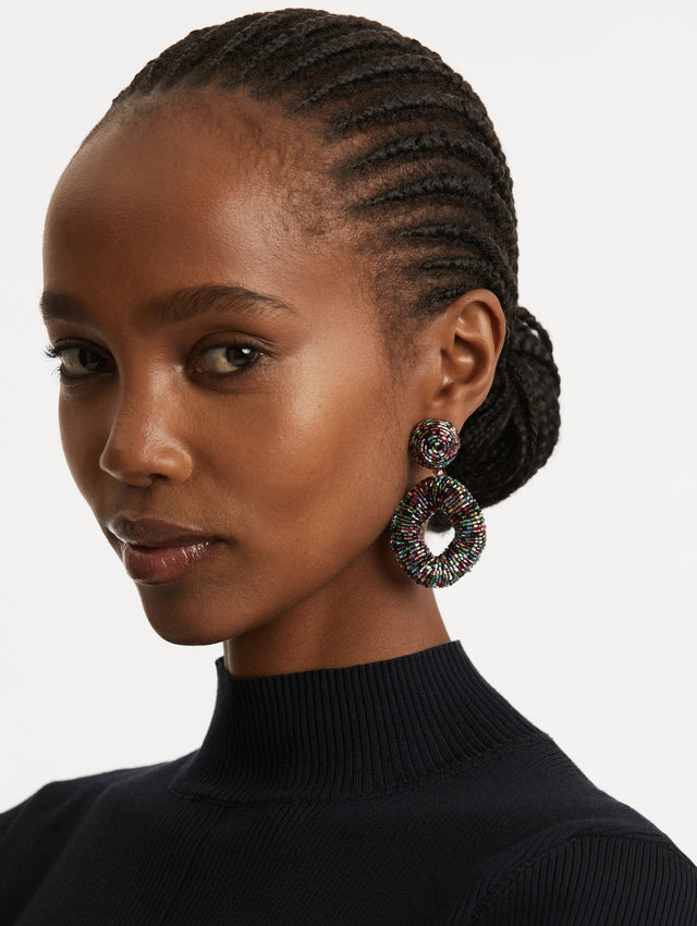 Model is wearing a hand-beaded pair of earrings which feature jet-black bugle beads in a bold, open-loop silhouette. 