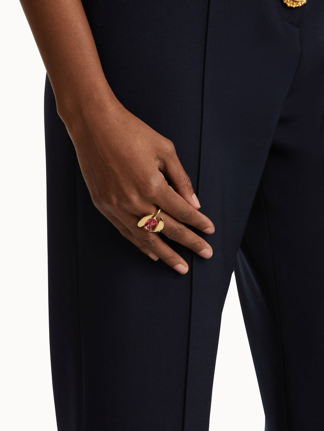 Model wearing a ring which frames a oval-cut stone in a curved setting. The polished gold-tone finish contrasts with the hue of the central gemstone. 