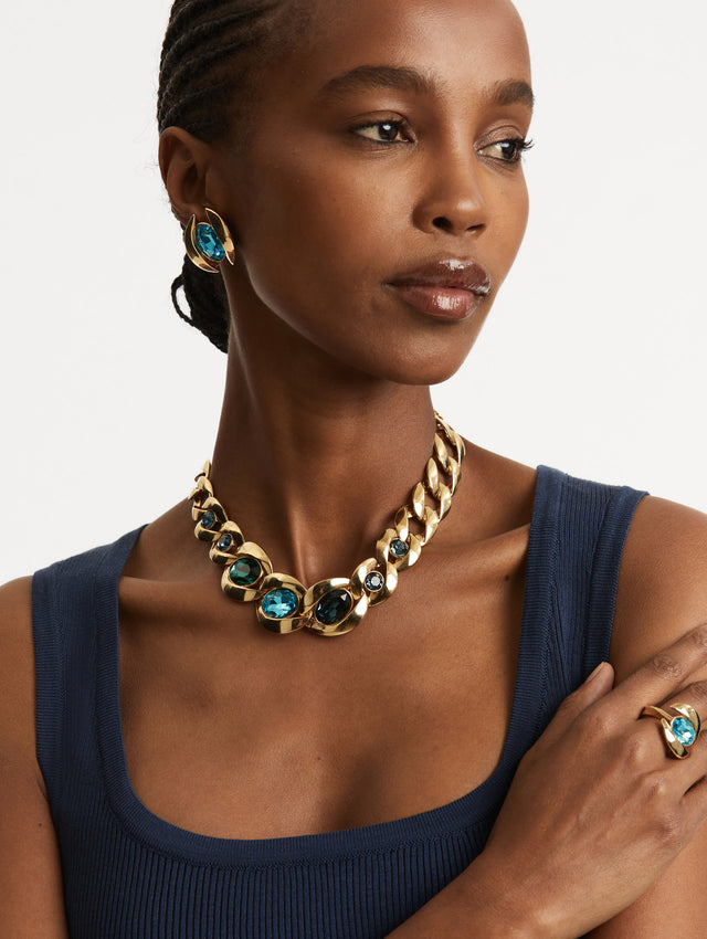 Model wearing a necklace with oversized polished links with a row of gemstones with sculpted bezels in varying cuts and blue hues—offering a contrast to the golden chain.