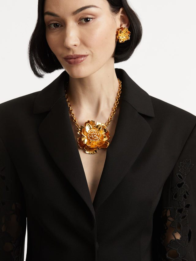 Model is wearing chain-link necklace featuring a peony pendant suspended at its center, cast in a gold finish with sculptural petals. A cluster of pavé crystals forms the middle of the bloom.