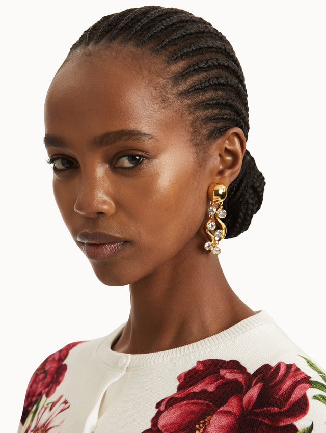 Model is wearing drop earrings that feature two polished gold-tone waves, each accented with brilliant round crystals, cascading from a sleek gold ball stud. 