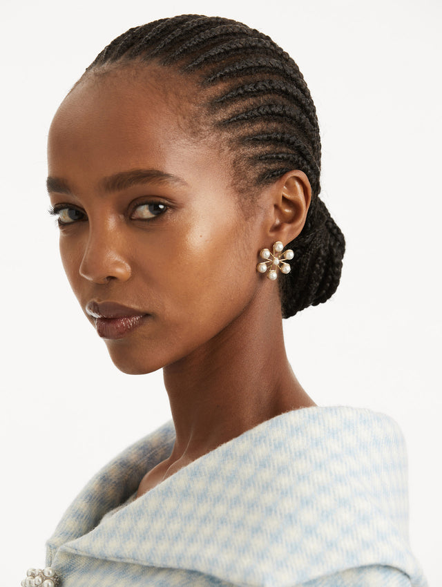 Model is wearing earrings that reimagine a flower from a central pearl, each petal is framed in polished gold and set with descending faux pearls. 