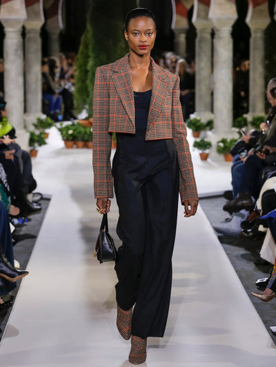 Holding a black top-handle handbag, the model walks the runway a cropped long-sleeve plaid tweed jacket, layered over a navy jumpsuit. Behind her, the blurred background reveals the audience and potted plants at the base of tall arches.
