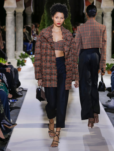 Holding a plaid gardenia appliqué handbag, the model walks the runway in a long-sleeve open-front plaid fringe jacket, layered over coordinating plaid cropped bustier top and blue trousers. Behind her, another model walks in the opposite direction, with the blurred background revealing the audience and potted plants at the base of tall arches.