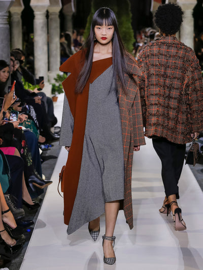 Holding a brown handbag, the model walks the runway in a long-sleeve open-front coat with a plaid and herringbone exterior, layered over a herringbone tweed dress. Behind her, another model walks in the opposite direction, with the blurred background revealing the audience and potted plants at the base of tall arches.