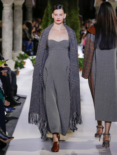The model walks the runway in an oversized open-front plaid fringe cardigan, layered over a herringbone tweed maxi dress. Behind her, another model walks in the opposite direction, with the blurred background revealing the audience and potted plants at the base of tall arches.
