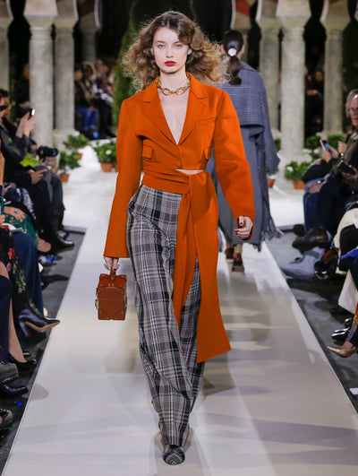 Holding an orange cube-like handbag, the model walks the runway in an orange long-sleeve cropped jacket with a tie-detail waist and plaid trousers. Behind her, another model walks in the opposite direction, with the blurred background revealing the audience and potted plants at the base of tall arches.