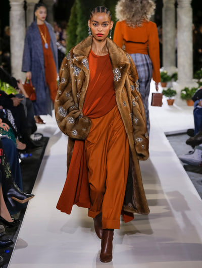 The model walks the runway in a brown open-front long-sleeve fur coat with crystal embroidery, layered over an oversized orange knit pullover and orange trousers. Behind her, several models walk the runway in various directions, with the blurred background revealing the audience and potted plants at the base of tall arches.