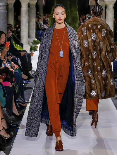 The model walks the runway in an open-front long-sleeve plaid coat, layered over an orange knit jumpsuit. Behind her, another model walks in the opposite direction, with the blurred background revealing the audience and potted plants at the base of tall arches.