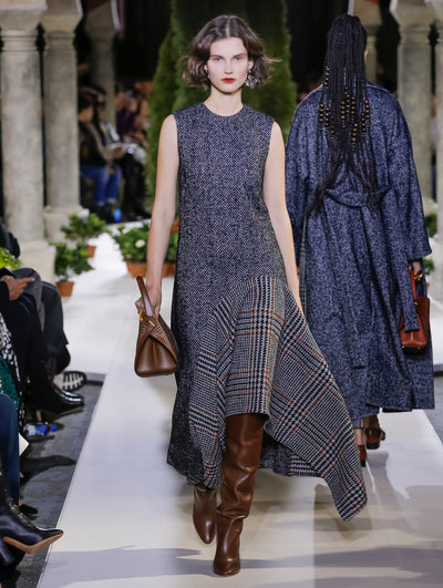 Holding a brown leather and plaid handbag, the model walks the runway in a herringbone tweed dress with a plaid inset on the skirt. Behind her, another model walks in the opposite direction, with the blurred background revealing the audience and potted plants at the base of tall arches.