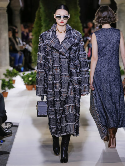 Holding a black cube-like handbag and wearing white sunglasses, the model walks the runway in a long-sleeve plaid and herringbone tweed patchwork fringe coat. Behind her, another model walks in the opposite direction, with the blurred background revealing the audience and potted plants at the base of tall arches.