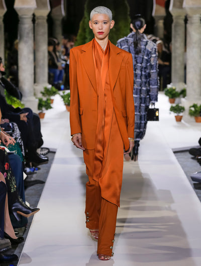 The model walks the runway in an orange long-sleeve double-breasted blazer, layered over an orange satin tie-neck blouse and orange trousers. Behind her, another model walks in the opposite direction, with the blurred background revealing the audience and potted plants at the base of tall arches.