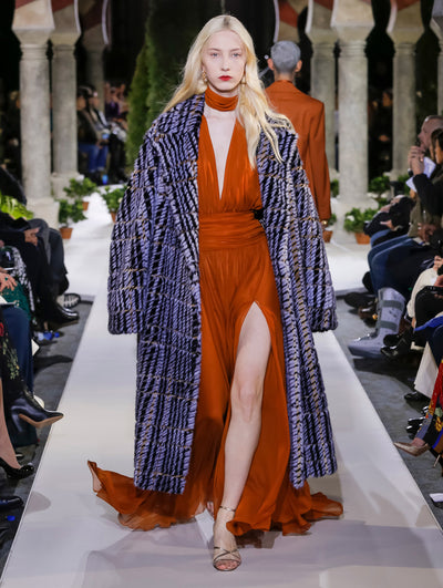 The model walks the runway in an open-front long-sleeve plaid fur coat, layered over an orange V-neck silk evening gown with scarf detail. Behind her, another model walks in the opposite direction, with the blurred background revealing the audience and potted plants at the base of tall arches.