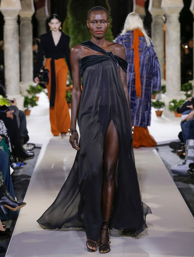 The model walks the runway in a black one-shoulder evening gown with a velvet bodice and silk skirt. Behind her, two models walk the runway in various directions, with the blurred background revealing the audience and potted plants at the base of tall arches.