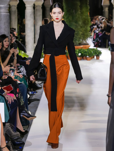 Holding a black top-handle handbag, the model walks the runway in a black long-sleeve jacket with sash detail and orange trousers. The blurred background reveals the audience and potted plants at the base of tall arches.