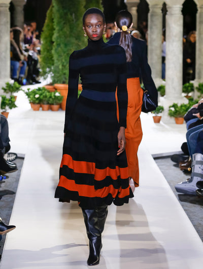 The model walks the runway in a black, navy and orange striped knit dress with long sleeves and a turtleneck. Behind her, another model walks in the opposite direction, with the blurred background revealing the audience and potted plants at the base of tall arches.
