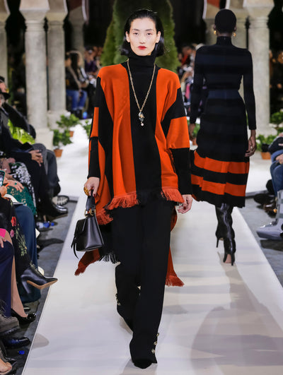 Holding a black top-handle handbag, the model walks the runway in a black and orange striped knit pullover with long sleeves, a turtleneck and fringe trim. Behind her, another model walks in the opposite direction, with the blurred background revealing the audience and potted plants at the base of tall arches.
