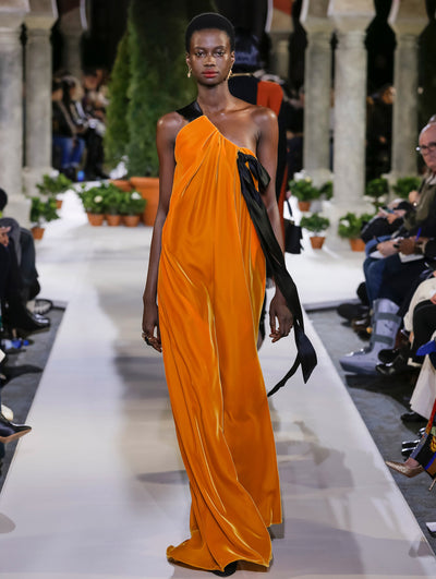 The model walks the runway in an orange one-shoulder bow-detailed evening gown. Behind her, another model walks in the opposite direction, with the blurred background revealing the audience and potted plants at the base of tall arches.