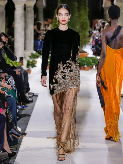 The model walks the runway in a black long-sleeve velvet cocktail dress with a threadwork embroidered tapestry design and floor-length fringe trim. Behind her, another model walks in the opposite direction, with the blurred background revealing the audience and potted plants at the base of tall arches.