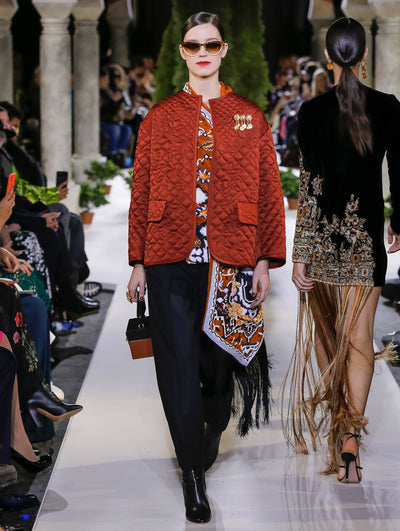 Holding a black and gold cube-like handbag and wearing brown sunglasses, the model walks the runway in a burnt orange bomber jacket with a brooch pinned to the chest, layered over a multi-colored knit blouse with fringe detailing and navy trousers. Behind her, another model walks in the opposite direction, with the blurred background revealing the audience and potted plants at the base of tall arches.