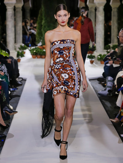 The model walks the runway in a strapless cocktail dress with a multi-colored tapestry design and fringe detailing. Behind her, another model walks in the opposite direction, with the blurred background revealing the audience and potted plants at the base of tall arches.