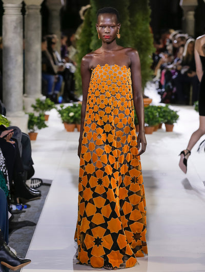 The model walks the runway in an orange strapless evening dress with a cutout geometric floral design. Behind her, another model walks in the opposite direction, with the blurred background revealing the audience and potted plants at the base of tall arches.