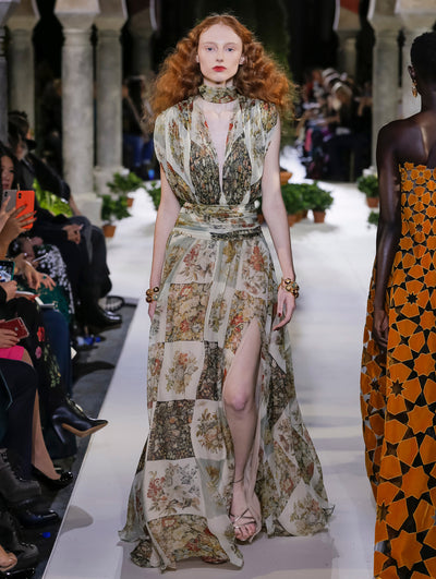 The model walks the runway in a sleeveless V-neck silk twill evening gown printed with a multi-colored floral patchwork design. Behind her, another model walks in the opposite direction, with the blurred background revealing the audience and potted plants at the base of tall arches.