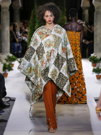 The model walks the runway in a zip-front oversized tunic printed with a multi-colored floral patchwork design, paired with orange trousers. Behind her, another model walks in the opposite direction, with the blurred background revealing the audience and potted plants at the base of tall arches.
