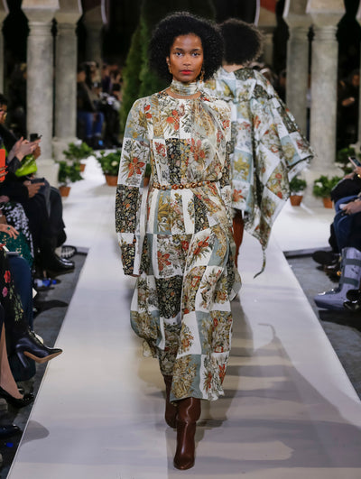 The model walks the runway in a long-sleeve silk twill evening gown featuring a multi-colored floral patchwork design with a self-belt and scarf detail. Behind her, another model walks in the opposite direction, with the blurred background revealing the audience and potted plants at the base of tall arches.