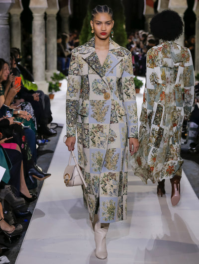 Holding an ivory shoulder bag, the model walks the runway in long-sleeve coat with a multi-colored floral patchwork design and gold button closure. Behind her, another model walks in the opposite direction, with the blurred background revealing the audience and potted plants at the base of tall arches.