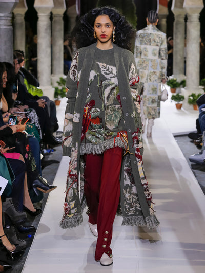 The model walks the runway in a gray oversized button-front knit cardigan with a multi-colored floral design, layered over a coordinating gray knit pullover with the same multi-colored floral design and burgundy trousers. Behind her, another model walks in the opposite direction, with the blurred background revealing the audience and potted plants at the base of tall arches.