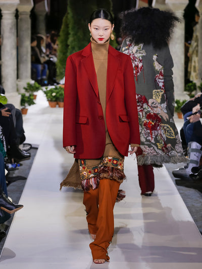The model walks the runway in a red long-sleeve double-breasted blazer, layered over a brown long-sleeve turtleneck knit pullover with floral embroidery and orange trousers. Behind her, another model walks in the opposite direction, with the blurred background revealing the audience and potted plants at the base of tall arches.