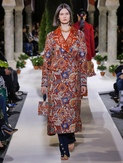 Holding a multi-colored cube-like handbag with a multi-colored floral design, the model walks the runway in a long-sleeve coat with the same floral design, layered over navy trousers. Behind her, another model walks in the opposite direction, with the blurred background revealing the audience and potted plants at the base of tall arches.