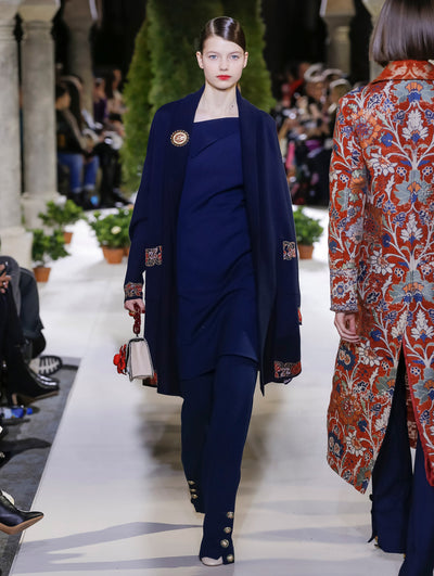 Holding an ivory and red gardenia appliqué handbag, the model walks the runway in a navy oversized open-front cardigan, layered over a navy tunic and navy trousers. Behind her, another model walks in the opposite direction, with the blurred background revealing the audience and potted plants at the base of tall arches.
