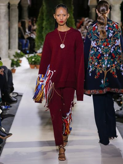The model walks the runway in a burgundy oversized long-sleeve knit pullover with multi-colored fringe detailing and burgundy trousers with the same fringe detailing. Behind her, another model walks in the opposite direction, with the blurred background revealing the audience and potted plants at the base of tall arches.