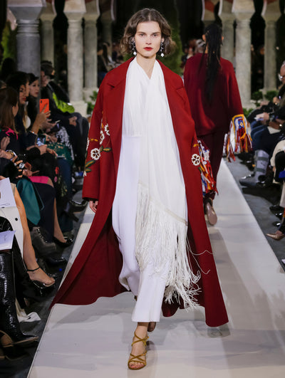 The model walks the runway in a red open-front long-sleeve wool coat with multi-color floral embroidery, layered over white jumpsuit with fringe detailing. Behind her, another model walks in the opposite direction, with the blurred background revealing the audience and potted plants at the base of tall arches.