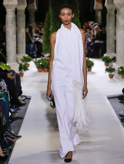 Holding a black and silver clutch, the model walks the runway in white sleeveless evening gown with fringe detailing. Behind her, the blurred background reveals the audience and potted plants at the base of tall arches.