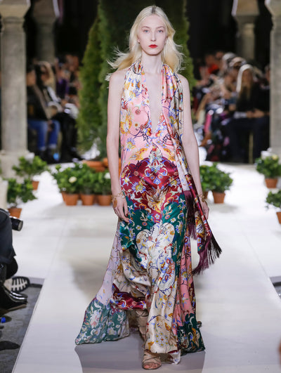 The model walks the runway in a sleeveless V-neck silk evening gown with a multi-colored tapestry design and fringe detailing. Behind her, the blurred background reveals the audience and potted plants at the base of tall arches.