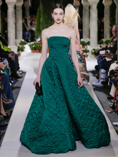 Holding a black clutch, the model walks the runway in a green strapless quilt evening gown. Behind her, another model walks in the opposite direction, with the blurred background revealing the audience and potted plants at the base of tall arches.