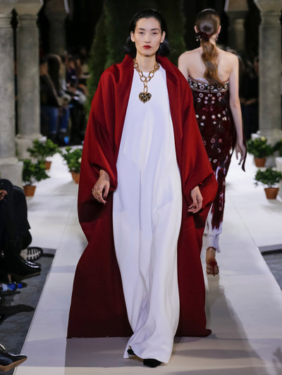 The model walks the runway in a burgundy long-sleeve open-front floor-length shawl, layered over a white column evening gown. Behind her, another model walks in the opposite direction, with the blurred background revealing the audience and potted plants at the base of tall arches.