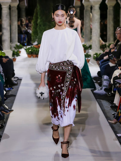 Holding a black and white sheep-shaped handbag, the model walks the runway in a white balloon sleeve blouse and burgundy wrap skirt with floral embroidery. Behind her, another model walks in the opposite direction, with the blurred background revealing the audience and potted plants at the base of tall arches.