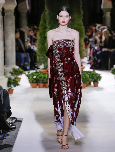 The model walks the runway in a burgundy strapless wrap evening dress with floral embroidery. Behind her, the blurred background reveals the audience and potted plants at the base of tall arches.