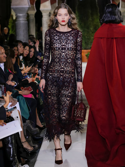 Holding a burgundy lamp-shaped handbag, the model walks the runway in a burgundy long-sleeve semi-sheer crochet dress with fringe trim. Behind her, another model walks in the opposite direction, with the blurred background revealing the audience and potted plants at the base of tall arches.