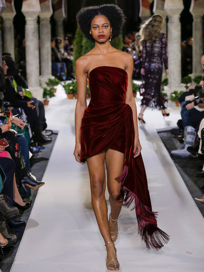 The model walks the runway in a burgundy strapless velvet cocktail dress with a draped bodice and fringe detailing. Behind her, another model walks in the opposite direction, with the blurred background revealing the audience and potted plants at the base of tall arches.
