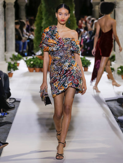 Holding a gardenia appliqué handbag with a multi-colored floral design, the model walks the runway in a one-shoulder cocktail dress with a draped bodice and the same floral design. Behind her, another model walks in the opposite direction, with the blurred background revealing the audience and potted plants at the base of tall arches.